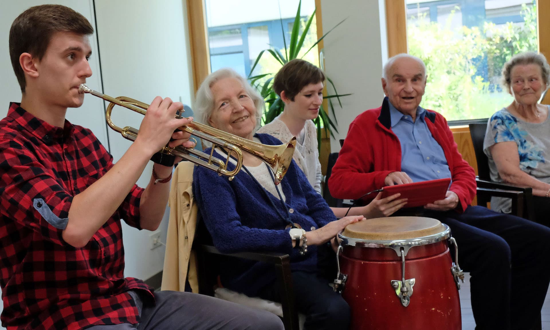 Ein junger Mann spielt Trompete, eine Seniorin trommelt auf einer Konga und lacht in die Kamera, eine junge Frau zeigt einem alten Mann ein Musikprogramm auf einem Tablet, eine alte Frau guckt interessiert zu.