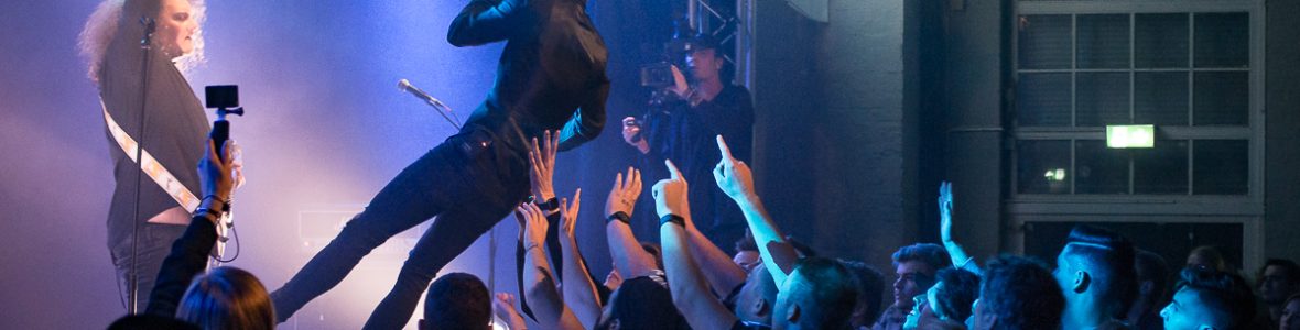 OLYMPUS DIGITAL CAMERA Ein junger Mann wirft sich im das Publikum zum Crowdsurfing, sein Bandmitglied steht ihm gegenüber auf der Bühne. Im Hintergrund sieht man einen Kameramann.